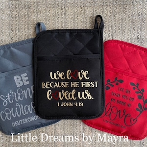 May include: Three quilted oven mitts with different colors and inspirational messages. The gray mitt says "Be strong and courageous, Deuteronomy 31:6". The black mitt says "We love because He first loved us, 1 John 4:19". The red mitt says "Let all that you do be done in love, 1 Corinthians 16:14".