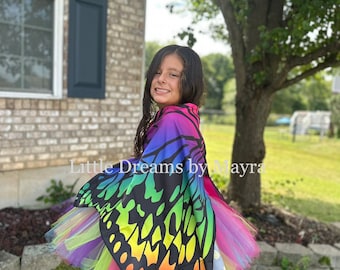Könnte beinhalten: Ein junges Mädchen trägt einen regenbogenfarbenen Tüll-Tutu-Rock und eine schwarze Schmetterlings-Kappe mit regenbogenfarbenen Flügeln. Auf der Kappe steht "Little Dreams by Mayra" gedruckt.
