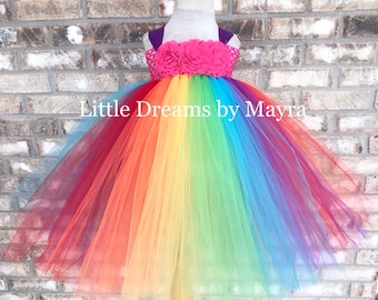 Puede incluir: Un vestido de tul de tutú arcoíris con un adorno de flores rosas en el corpiño. El vestido está en un maniquí de madera y tiene el texto "Little Dreams by Mayra" en la falda.