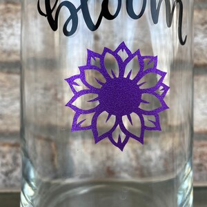 Op de afbeelding: Een heldere glazen fles met een paarse glitter-zonnebloem sticker en het woord "bloom" in zwarte cursieve letters.