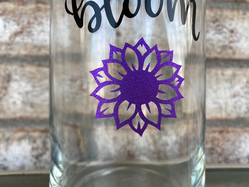 Op de afbeelding: Een heldere glazen fles met een paarse glitter-zonnebloem sticker en het woord "bloom" in zwarte cursieve letters.