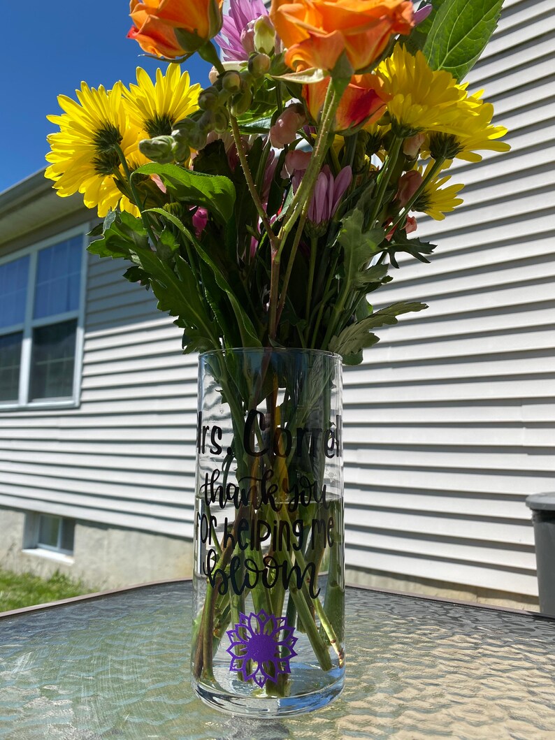 Op de afbeelding: Een heldere glazen vaas met een paarse zonnebloem sticker en de tekst "Mrs. Cornell thank you for helping me bloom" in zwarte letters. De vaas is gevuld met een boeket gele, oranje en paarse bloemen.