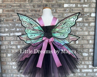 Puede incluir: Tutu de tul negro con alas de mariposa iridiscentes y un lazo de satén rosa. El tutu está en un soporte de madera. "Little Dreams by Mayra" está escrito en el tutu.