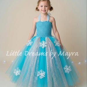 May include: A floor-length turquoise tulle dress featuring a crocheted bodice, halter-style straps, and a full skirt layered with shimmering silver tulle panels. The dress is decorated with white snowflake appliqués and includes the text Little Dreams by Mayra.