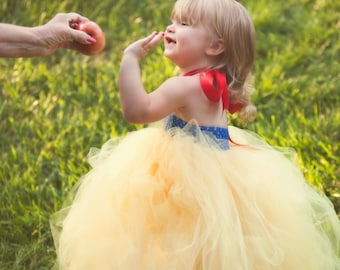 Puede incluir: Una niña pequeña vestida de Blancanieves, con un vestido de tul amarillo, un corpiño azul y un lazo rojo, está de pie en un campo de hierba y extiende la mano hacia una manzana roja.