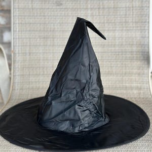 May include: A black pointed hat with a wide, circular brim. The material of the hat has a wrinkled, slightly glossy texture, and the top of the cone is bent over to one side. The hat is constructed from a lightweight, flexible fabric material.