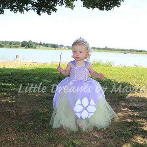 May include: A young child wearing a lavender and green tutu dress with a silver tiara and holding a stick. The dress has a bodice with a beaded design and a large white flower-like shape on the skirt. The text "Little Dreams by Mayra" is visible.