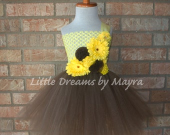 May include: A brown tulle tutu with a yellow and brown flower embellishment. The bodice is a yellow crochet fabric with a single yellow strap. The text "Little Dreams by Mayra" is visible on the skirt.