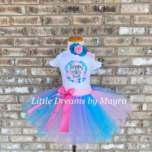 May include: A white baby onesie with the text "Happy Easter Girl" in a floral wreath design. The onesie is paired with a blue and pink tulle tutu skirt with a pink satin ribbon.