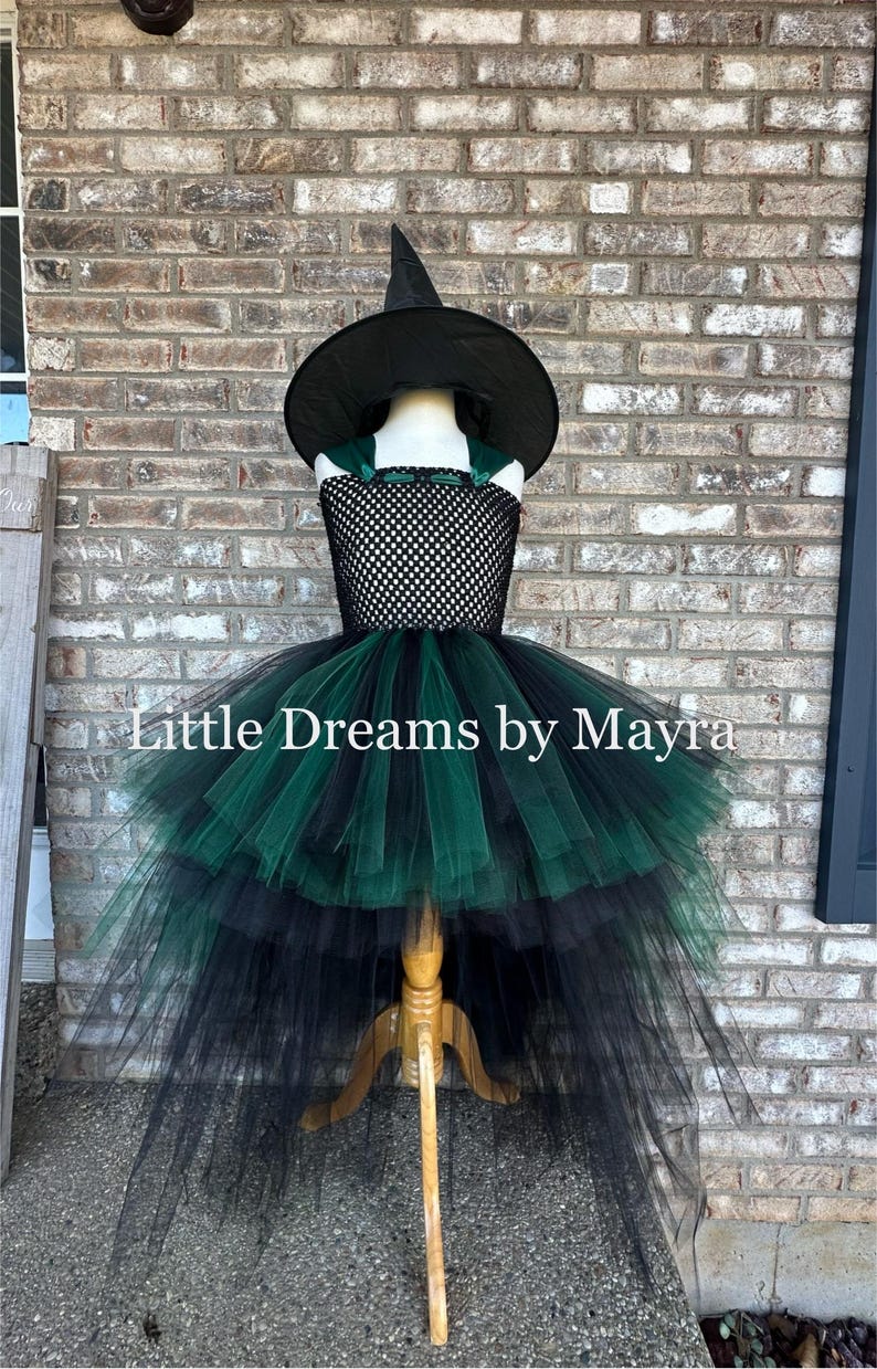 May include: A black witch hat and a dress featuring a black crocheted bodice with a high-low skirt made of layered black and dark green tulle. The text Little Dreams by Mayra is centered across the middle of the dress in white lettering.