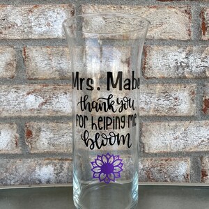 Op de afbeelding: Een heldere glazen vaas met een zwarte vinyl sticker met de tekst "Mrs. Mable thank you for helping me bloom". Een paarse bloemgrafiek bevindt zich onder de tekst. De vaas staat op een bakstenen oppervlak.