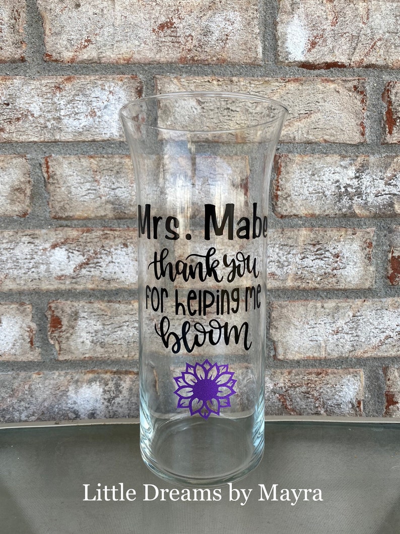 Op de afbeelding: Een heldere glazen vaas met een zwarte vinyl sticker met de tekst "Mrs. Mable thank you for helping me bloom". Een paarse bloemgrafiek bevindt zich onder de tekst. De vaas staat op een bakstenen oppervlak.