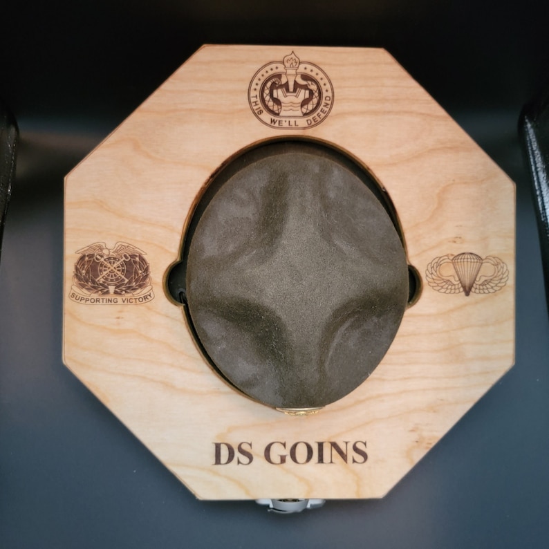 May include: A wooden hat display with a green hat, the text "DS GOINS" and three military insignia. The insignia are "This We'll Defend", "Supporting Victory", and a parachute.
