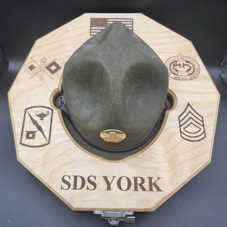 Puede incluir: Un expositor de sombreros de madera con un sombrero militar verde, una bandera estadounidense y el texto "SDS YORK".