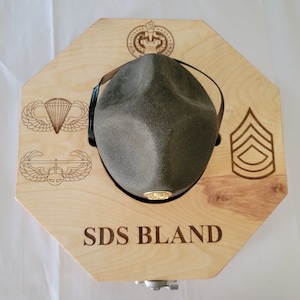 May include: A wooden hat display with a green hat, a gold pin, and a name engraved on the front. The display also features three military insignia: a parachute, a helicopter, and a rank insignia.