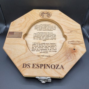 May include: A wooden plaque with a hexagonal shape and a cutout in the center. The plaque is engraved with the text "I am a Drill Sergeant" and "DS ESPINOZA". The text is in a serif font and is centered on the plaque. The plaque is a light brown color and has a natural wood grain.