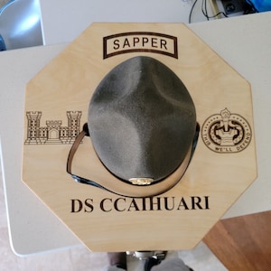May include: A wooden hat display with a green military hat, the words "SAPPER" and "DS CCAIHUARI" are engraved on the display, along with a military insignia and a castle.