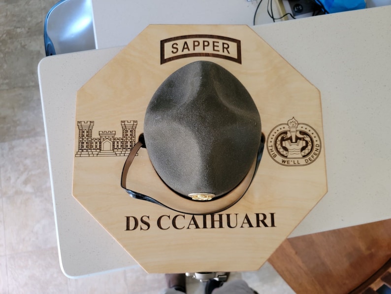 May include: A wooden hat display with a green military hat, the words "SAPPER" and "DS CCAIHUARI" are engraved on the display, along with a military insignia and a castle.