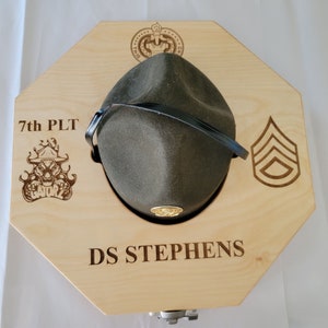 May include: A wooden hat rack with a green hat, a black strap, and a gold emblem. The rack is engraved with "7th PLT", a skull with flames, "DS STEPHENS", and a chevron.