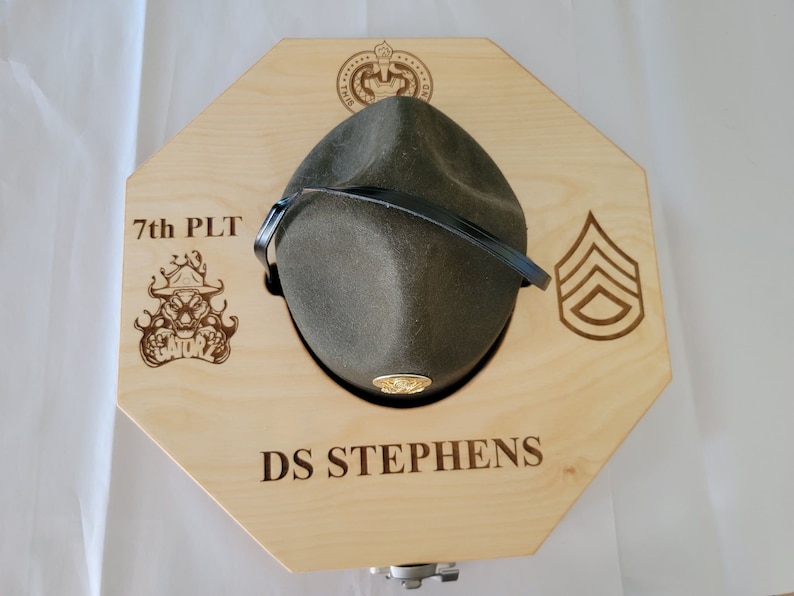 May include: A wooden hat rack with a green hat, a black strap, and a gold emblem. The rack is engraved with "7th PLT", a skull with flames, "DS STEPHENS", and a chevron.