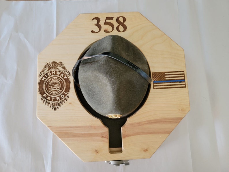 May include: A wooden hat rack with a hat on it. The rack is octagonal and has a blue line American flag and a Highway Patrol badge engraved on it. The number 358 is also engraved on the rack.