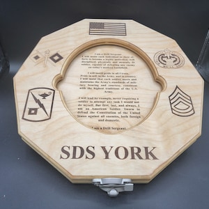 Puede incluir: Una placa de madera con una cita sobre los deberes de un sargento de instrucci&oacute;n en el ej&eacute;rcito de los Estados Unidos. La placa es octogonal y presenta una variedad de insignias militares, incluido el logotipo del ej&eacute;rcito de los Estados Unidos, un rayo y una l&aacute;mpara, y un gal&oacute;n. El texto dice: "Soy un sargento de instrucci&oacute;n... Dar&eacute; el ejemplo, nunca pidiendo a un soldado que intente una tarea que yo no har&iacute;a. Pero primero, &uacute;ltimo y siempre, soy un soldado estadounidense. Jurado para defender la Constituci&oacute;n de los Estados Unidos contra todos los enemigos, tanto extranjeros como nacionales. Soy un sargento de instrucci&oacute;n." La placa est&aacute; firmada "SDS YORK".