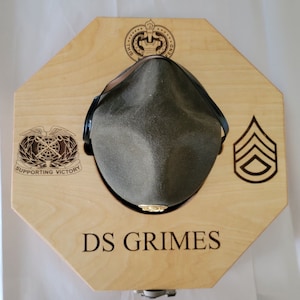 May include: A wooden hat display stand with an octagonal shape. The stand has a cutout for a hat and features a military insignia, a rank insignia, and the text "DS GRIMES".