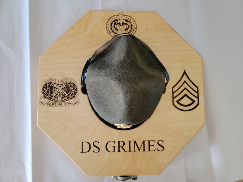 May include: A wooden hat display stand with an octagonal shape. The stand has a cutout for a hat and features a military insignia, a rank insignia, and the text "DS GRIMES".