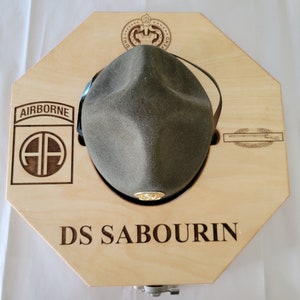 May include: A wooden hat display box with an octagonal shape. The box has a cutout for a hat and a leather strap to hold the hat in place. The box is engraved with the words "Airborne", "AA", "DS SABOURIN", and a military insignia.
