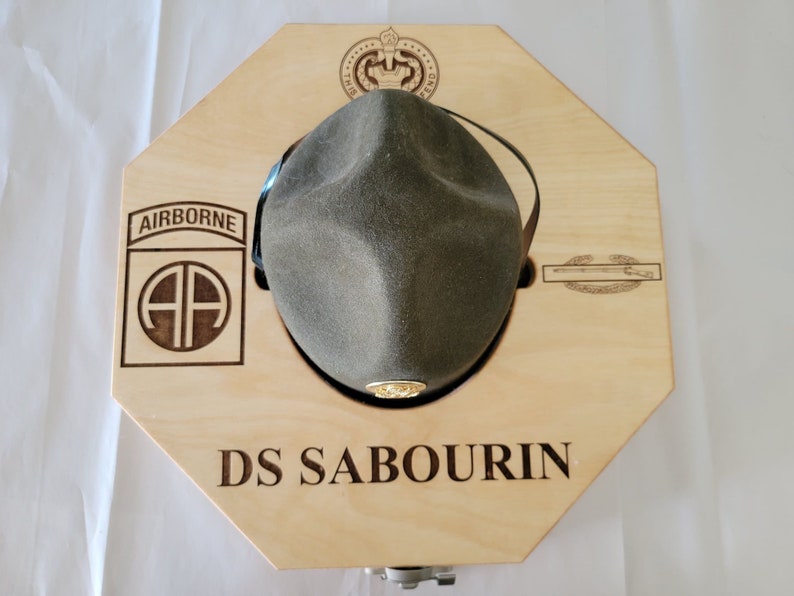 May include: A wooden hat display box with an octagonal shape. The box has a cutout for a hat and a leather strap to hold the hat in place. The box is engraved with the words "Airborne", "AA", "DS SABOURIN", and a military insignia.