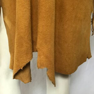 May include: A tan suede fringed vest. The vest has a draped, open front design and long fringe detailing on the sleeves. The suede material has a textured appearance. The vest is displayed on a mannequin.