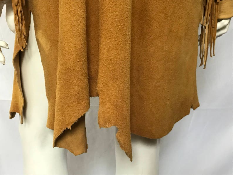 May include: A tan suede fringed vest. The vest has a draped, open front design and long fringe detailing on the sleeves. The suede material has a textured appearance. The vest is displayed on a mannequin.