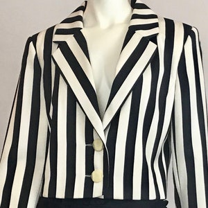 Puede incluir: Una chaqueta de rayas blancas y negras con cuello de solapa y cierre de botones. La chaqueta tiene mangas largas y una longitud recortada.
