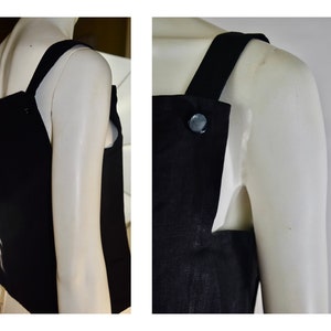 May include: A black sleeveless top with wide straps and a large button detail. The top is displayed on a mannequin, showcasing its simple yet elegant design. The fabric appears to be a solid, dark color.