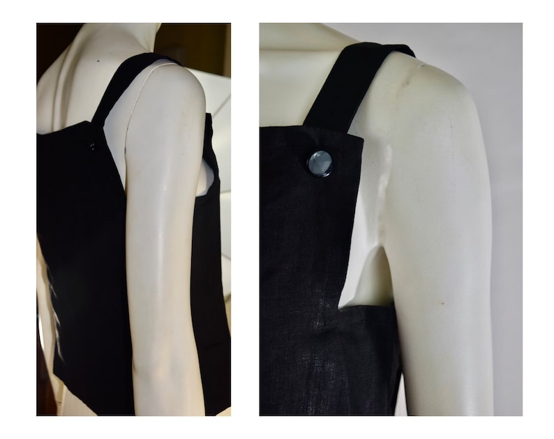 May include: A black sleeveless top with wide straps and a large button detail. The top is displayed on a mannequin, showcasing its simple yet elegant design. The fabric appears to be a solid, dark color.