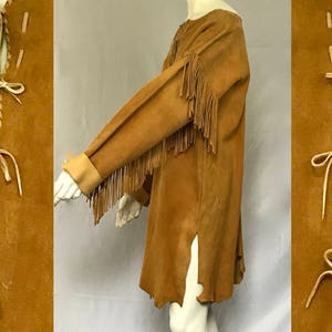 May include: A tan suede tunic with long sleeves and fringe detail on the sleeves. The tunic has a split on the side and is displayed on a mannequin. The side panels feature decorative stitching.
