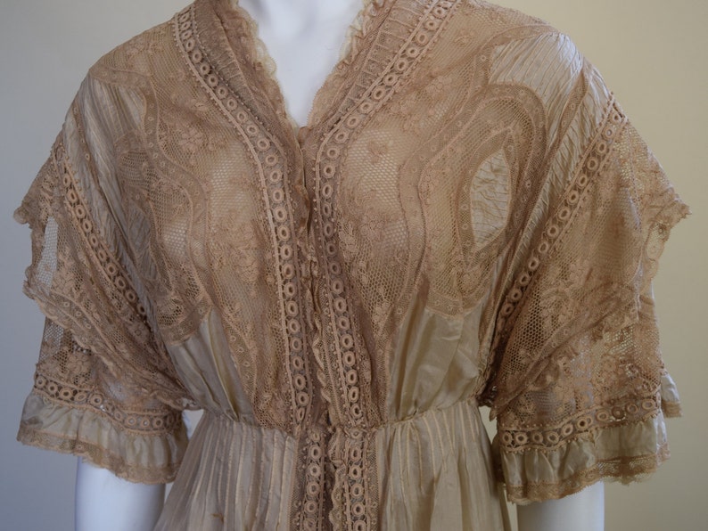 May include: A vintage beige dress with intricate lace detailing. The dress features a V-neckline, short sleeves, and a fitted waist. The lace is used on the bodice, sleeves, and along the button-down front. The skirt is pleated and flows down from the waist.