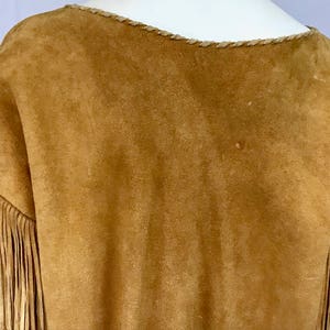 May include: A tan suede top with fringe detail on the sleeves. The neckline is trimmed with a braided design. The garment has a soft, textured appearance. The overall style suggests a bohemian or Western-inspired aesthetic.