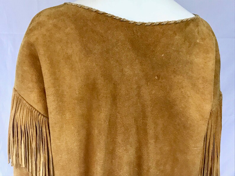 May include: A tan suede top with fringe detail on the sleeves. The neckline is trimmed with a braided design. The garment has a soft, textured appearance. The overall style suggests a bohemian or Western-inspired aesthetic.