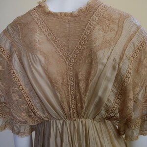 May include: A vintage cream-colored dress with intricate lace detailing. The dress features a high neckline, a fitted bodice, and a flowing skirt. The lace is a light tan color and is used on the sleeves and bodice.
