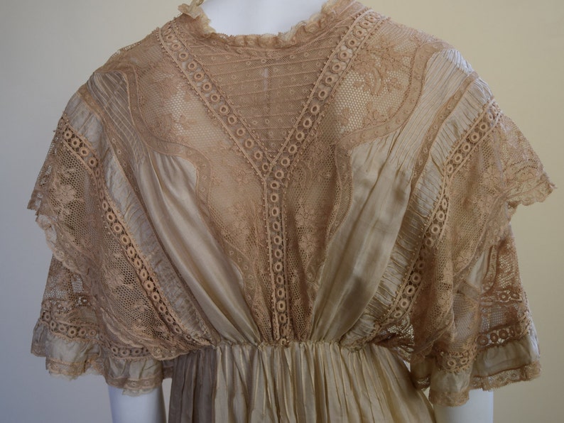 May include: A vintage cream-colored dress with intricate lace detailing. The dress features a high neckline, a fitted bodice, and a flowing skirt. The lace is a light tan color and is used on the sleeves and bodice.