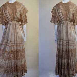 May include: A vintage beige dress with intricate lace detailing. The dress features a high neckline, short sleeves, and a flowing skirt with multiple lace-trimmed tiers. The dress is displayed on a mannequin, showcasing its elegant design and vintage aesthetic.