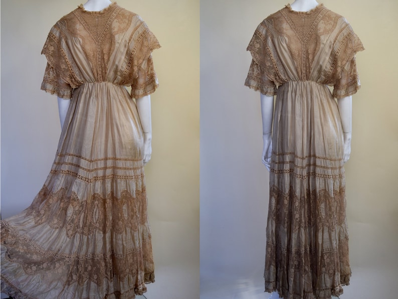 May include: A vintage beige dress with intricate lace detailing. The dress features a high neckline, short sleeves, and a flowing skirt with multiple lace-trimmed tiers. The dress is displayed on a mannequin, showcasing its elegant design and vintage aesthetic.