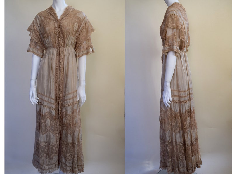 May include: A vintage beige dress with intricate lace detailing. The dress features a V-neckline, short sleeves, and a floor-length skirt. The lace trim is a darker shade of brown, creating a contrast against the lighter fabric. The dress is displayed on a mannequin.