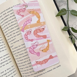 May include: Pink and orange shrimp illustrations on a bookmark with a white background and pink and orange stripes. The bookmark has a pink and orange string attached to the top.