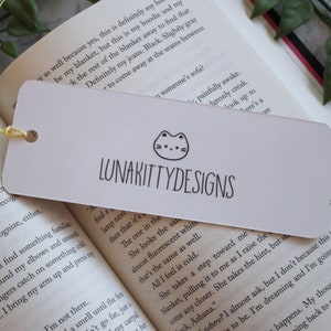 May include: White bookmark with a black and white illustration of a cat with a pink nose and a black outline. The text "LUNAKITTYDESIGNS" is printed in black below the cat illustration.