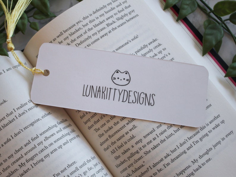 May include: White bookmark with a black and white illustration of a cat with a pink nose and a black outline. The text "LUNAKITTYDESIGNS" is printed in black below the cat illustration.