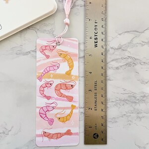 May include: A pink and yellow shrimp bookmark with a pink tassel. The bookmark is about 6 inches long and features a watercolor-style background with pink and white stripes.