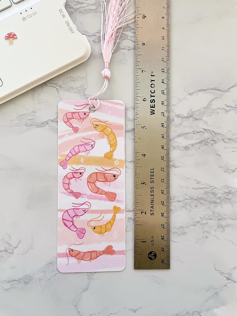 May include: A pink and yellow shrimp bookmark with a pink tassel. The bookmark is about 6 inches long and features a watercolor-style background with pink and white stripes.