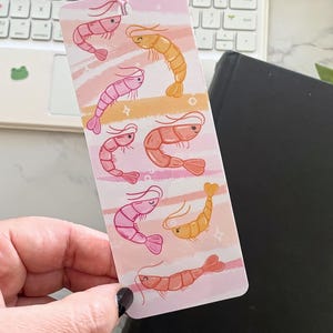 May include: A pink and orange bookmark with a whimsical design of multiple shrimp. The shrimp are illustrated in a cartoon style with different expressions and are set against a background of pink and orange stripes.